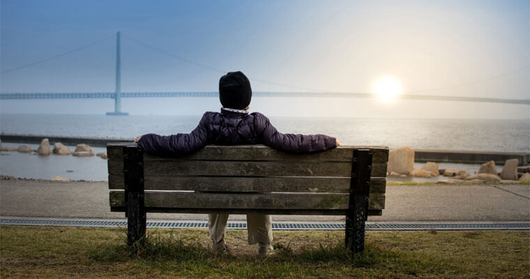 A person sits on a bench and watches the sun set behind a suspension bridge.